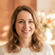 Smiling German woman in her late 30s, light brown hair, friendly expression, blurred warm amber and peach background.